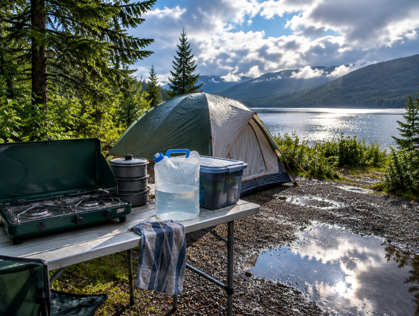 A half-packed camp kitchen table beside a rain-beaded tent at a lakeside campsite as the sun breaks through.