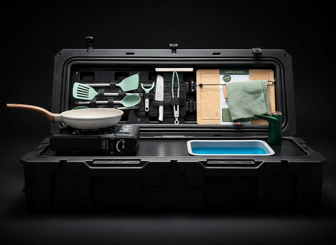 Portable kitchen setup with cookware, utensils, and a hot plate inside a black case on a dark background.