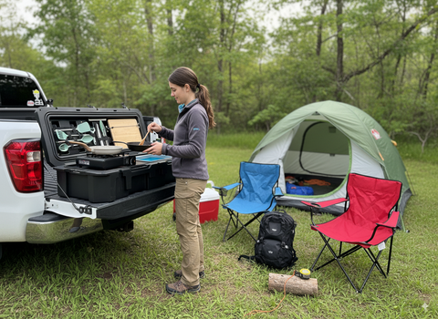 Person preparing food in a truck bed with camping kitchen and a tent in the background