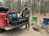 Man setting up camp kitchen in a truck bed with chairs and a duffel bag nearby.